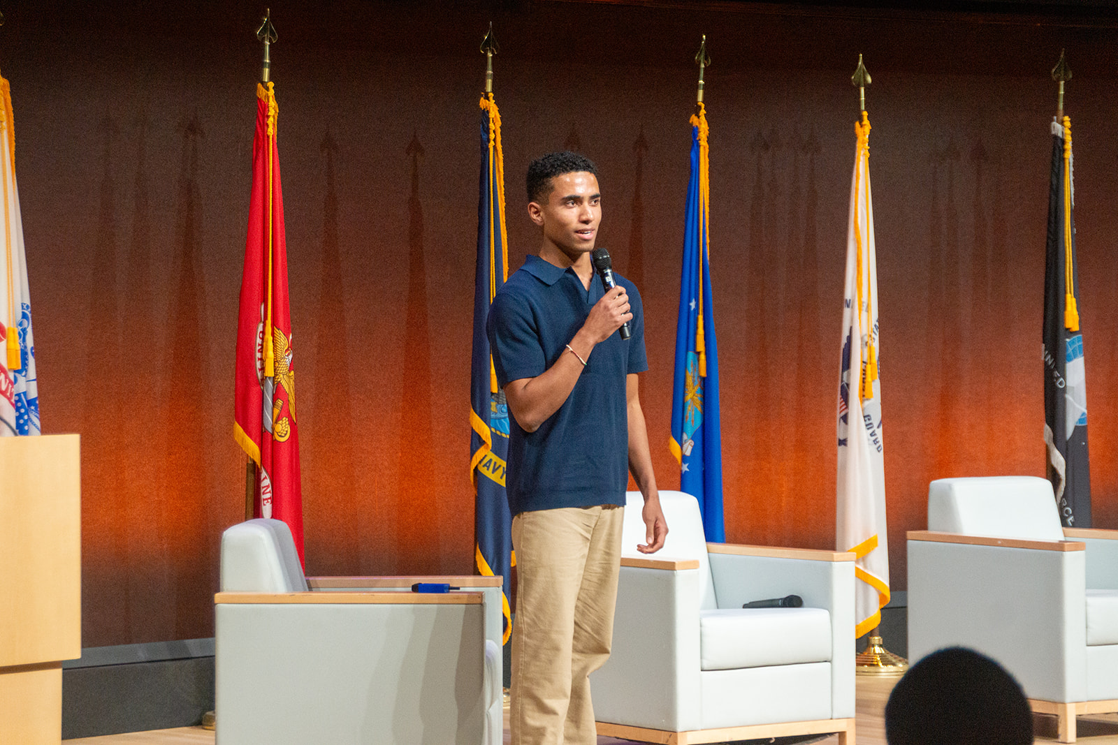Orion Goodman speaking with a microphone on stage in front of a row of flags.